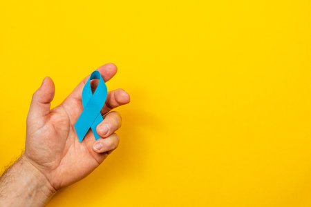 A male hand grips a blue ribbon, representing awareness and support for cancer detection and treatment. The bright yellow background highlights the importance of the cause.の写真素材