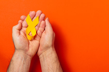 Two hands gently cradle a yellow ribbon, representing solidarity and support in the fight against cancer. The vibrant red background enhances the message of awareness and care.の写真素材