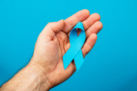 In a vibrant blue background, a male hand holds a blue ribbon, representing awareness for cancer. This powerful symbol emphasizes the importance of detection and support for patients.の写真素材