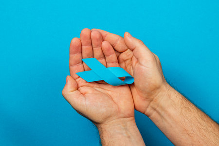 A pair of hands hold a blue ribbon against a bright blue background, symbolizing support for cancer awareness and prevention efforts. The image emphasizes courage and hope.の写真素材