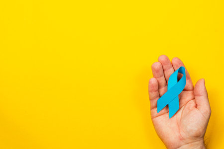 A male hand holds a blue ribbon against a bright yellow background, symbolizing awareness and support for cancer detection and treatment efforts.の写真素材
