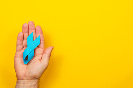 A male hand holds a blue awareness ribbon against a bright yellow background. This symbolizes support and awareness for prostate cancer and related health issues.の写真素材