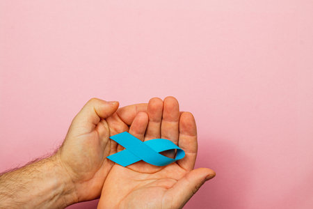 A pair of hands gently cradle a blue cancer awareness ribbon against a soft pink background. This symbolizes hope, support, and the ongoing fight against cancer.の写真素材