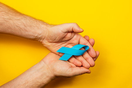 Two hands hold a blue ribbon against a vibrant yellow backdrop, symbolizing support for cancer awareness and highlighting the importance of early detection and treatment.の写真素材