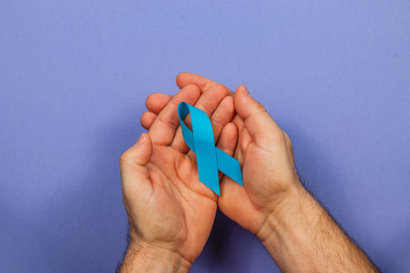 Hands hold a blue ribbon against a solid background, symbolizing awareness and support for cancer detection initiatives. This visual highlights the importance of cancer care and prevention.の写真素材