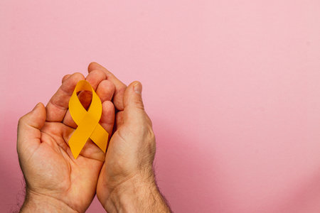 A pair of hands gently holds a yellow ribbon against a soft pink background, representing awareness and solidarity in the fight against cancer and promoting health support.の写真素材