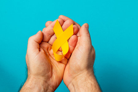 Two hands hold a yellow ribbon against a blue background, representing support, awareness, and courage in the battle against cancer and its challenges.の写真素材