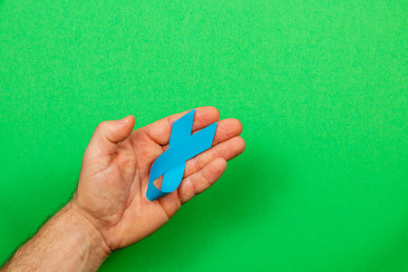 A male hand holds a blue ribbon symbolizing cancer awareness against a bright green background. This representation emphasizes the importance of early detection and support for patients.の写真素材