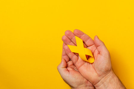 A pair of hands tenderly hold a yellow ribbon, symbolizing hope and awareness for cancer. The bright yellow background enhances the message of solidarity in the fight against illness.の写真素材