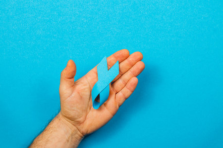 A male hand holds a blue ribbon against a bright blue background, symbolizing cancer awareness. This act raises support for patients and highlights the importance of detection and care.の写真素材