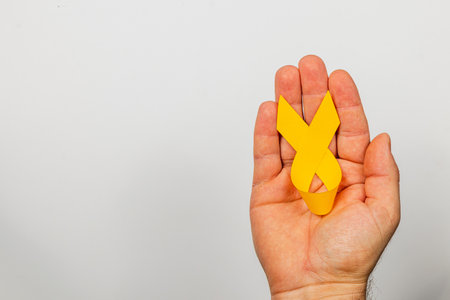A woman holds a yellow ribbon in her hand against a plain background, representing courage and solidarity in the fight against cancer and promoting awareness for early detection and support.の写真素材