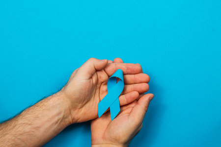 A person holds a blue ribbon between their hands against a solid blue background. This gesture signifies support for cancer awareness and highlights the importance of examination and early detection.の写真素材