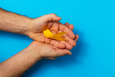 Two hands gently cradle a yellow ribbon, representing solidarity and hope for cancer awareness, treatment, and the fight against illness, while set against a bright blue background.の写真素材