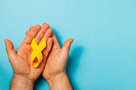 A person holds a yellow ribbon gently in their hands against a bright blue background, symbolizing hope and support for those battling cancer and raising awareness of the illness.の写真素材