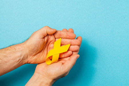 Hands hold a yellow ribbon against a light blue background, symbolizing support for cancer awareness and prevention. This represents hope, courage, and solidarity in battling the disease.の写真素材