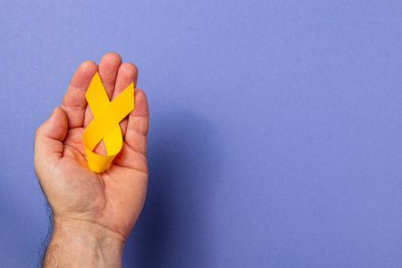 A hand holds a yellow ribbon representing cancer awareness. The background is a solid light blue, emphasizing the importance of support and solidarity in the fight against illness.の写真素材