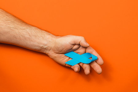 A male hand holds a symbolic blue ribbon against a vibrant orange backdrop. This represents support for cancer awareness and the fight against various cancer types.の写真素材