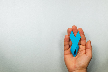 A male hand holds a blue awareness ribbon against a light grey background. The ribbon represents support for cancer detection, treatment, and survival. This symbolizes courage and hope.の写真素材