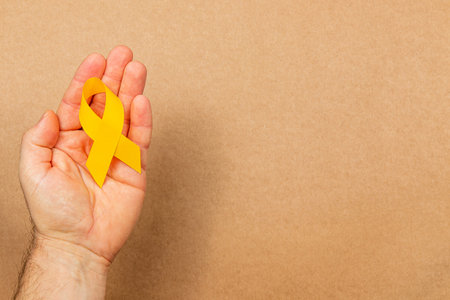 A hand holds a yellow ribbon, symbolizing awareness for cancer screening and support for those affected by illness. The background is a neutral color, enhancing focus on the ribbon.の写真素材