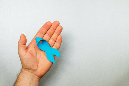 A male hand holds a vibrant blue ribbon against a neutral background, representing dedication to cancer awareness and prevention efforts.の写真素材