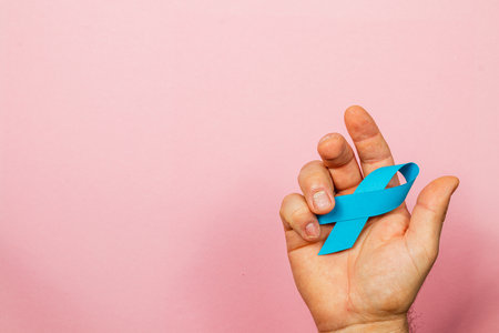 A male hand holds a blue ribbon against a soft pink background, symbolizing awareness and support for cancer detection and treatment information.の写真素材