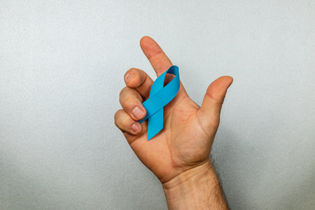 A male hand holds a blue ribbon, representing support for cancer awareness. The background is grey, emphasizing the ribbonâs color. This visual promotes awareness and care for cancer patients.の写真素材