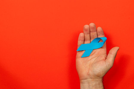 A male hand holds a blue ribbon against a vibrant red background, symbolizing cancer awareness and support for treatment and prevention efforts related to various cancers.の写真素材
