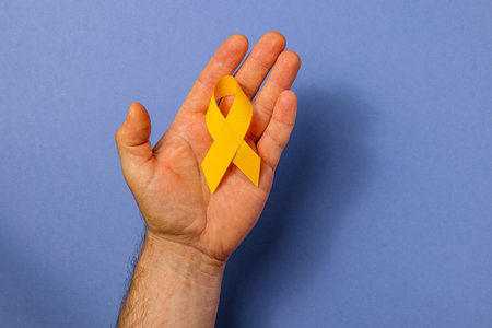 A person's hand holds a yellow ribbon against a solid color background, symbolizing cancer awareness.の写真素材