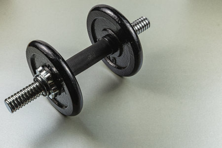 A black metal dumbbell rests on a smooth gray surface, highlighting its sturdy design and suitability for strength training and exercise routines.の写真素材