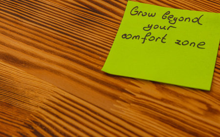 A bright green sticky note with the message to grow beyond your comfort zone rests on a wooden...