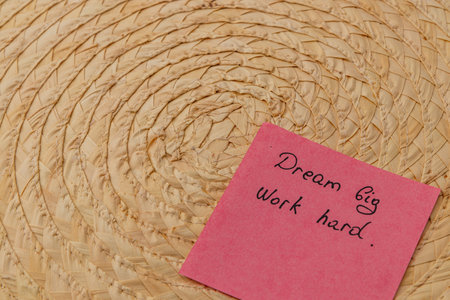 A pink sticky note featuring motivational phrases sits next to a round woven basket. The focus highlights the text encouraging hard work and big dreams, suggesting a positive mindset.の写真素材