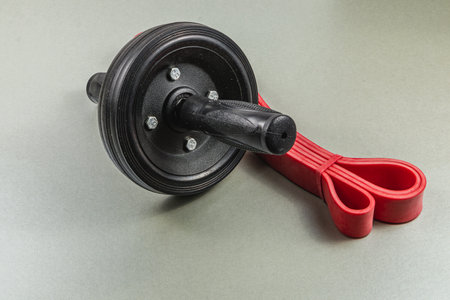 A black ab roller with a sturdy handle sits beside a red resistance band on a gray surface. These tools are perfect for enhancing core strength during workouts.の写真素材