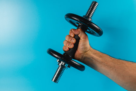 A hand grips a black dumbbell with silver ends, showcasing strength and focus. The bright blue background emphasizes the activity and determination.の写真素材
