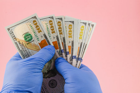 A pair of gloved hands displays a fan of hundred dollar bills against a pink background, illustrating currency handling and the importance of hygiene.の写真素材