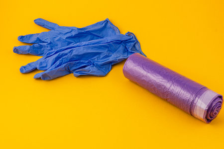 A pair of blue latex gloves lies next to a roll of purple trash bags on a bright yellow surface. This arrangement highlights a clean and organized approach to waste management.の写真素材