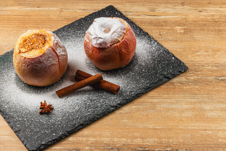 Two baked apples dusted with powdered sugar sit on a dark slate tray. Nearby, two cinnamon sticks and a star anise add to the cozy autumn atmosphere.の写真素材