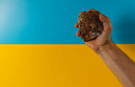 A hand firmly grips a rugged stone, contrasting against a bright blue and yellow backdrop, symbolizing resilience and strength in a vibrant setting.の写真素材