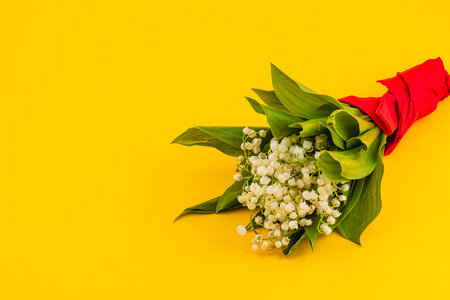 A vibrant bouquet of lilies of the valley wrapped in bright red paper rests on a sunny yellow surface, creating a cheerful and striking display of nature's beauty.の写真素材