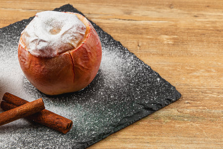 A baked apple topped with powdered sugar sits on a dark slate board. Cinnamon sticks are placed nearby, creating a cozy atmosphere perfect for fall.の写真素材