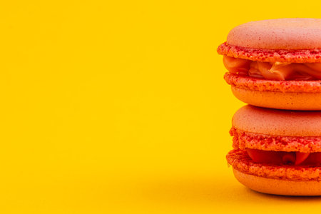 Two vibrant orange macarons rest on top of each other, showcasing their delicate texture and creamy filling against a cheerful yellow background.の写真素材