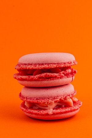 Two layers of pink macarons filled with cream stand tall against a striking orange backdrop, showcasing their delightful texture and color contrast.の写真素材
