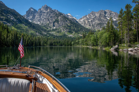 A calm lake mirrors an American flag as a boat glides through the water, framed by majestic mountains and lush green forests under a clear blue sky.の素材