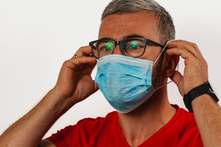 A man with glasses and short hair is securing a blue face mask on his face, preparing for safe...