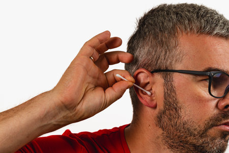 A man gently cleans his ear with a cotton swab while seated indoors, focusing on personal hygiene in...
