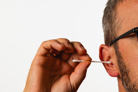 A man uses a cotton swab to clean his ear while sitting indoors during daylight. The background is...