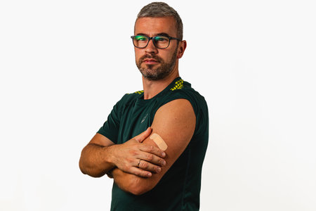 A man stands confidently with his arms crossed, displaying a bandage on his arm. He is in an indoor...