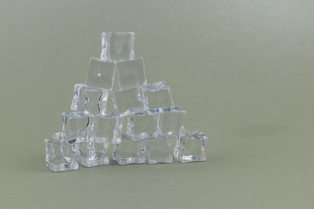 Many clear ice cubes are stacked in a pyramid formation on a smooth, neutral-colored surface, creating a visually appealing display.の写真素材