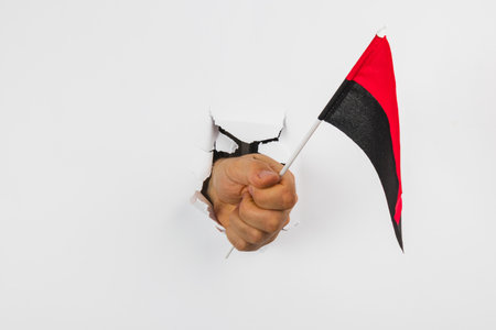 A fist emerges through a white wall gripping a small black and red flag, symbolizing strength and determination in a bold display.の写真素材