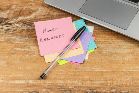 Colorful sticky notes labeled with human resources tasks sit next to a pen and a laptop on a wooden desk, suggesting office organization and planning.の写真素材