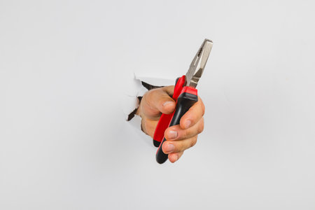A hand emerges from a hole in a white wall, firmly grasping a pair of pliers. This scene showcases a surprising moment, emphasizing the tool's details.の写真素材